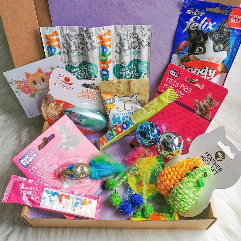 May include: A cardboard box filled with cat toys and treats. The box contains a variety of items, including catnip, a laser pointer, a feather toy, a catnip toy, a catnip mouse, and a catnip stick. The box also includes a card with a cat on it and a bag of cat food.