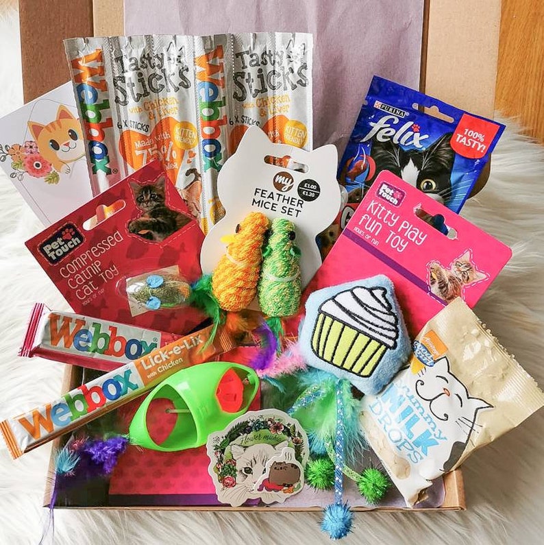 May include: A cat gift box filled with various cat toys, treats, and catnip. The box includes a variety of items, such as a feather mouse toy, a cupcake-shaped toy, a catnip toy, and a variety of cat treats. The box is decorated with a pink and purple theme.