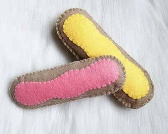 Puede incluir: Dos eclairs de fieltro, uno con relleno rosa y otro con relleno amarillo. Los eclairs son marrones con una puntada blanca alrededor de los bordes.