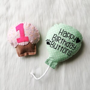 May include: A green felt balloon with white polka dots and the text "Happy Birthday Buttons" in black. A pink felt cupcake with a number one and sprinkles is next to the balloon.