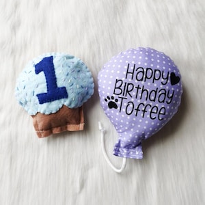 May include: A purple balloon with white polka dots and the text "Happy Birthday Toffee" with a paw print and a heart. A blue cupcake with white sprinkles and the number "1" in blue.