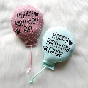 Two fabric catnip cat toy balloons with polka dots and strings. The pink balloon says 'Happy Birthday Fifi' with a paw print and a heart. The blue balloon says 'Happy Birthday Ginge' with a paw print and a heart.