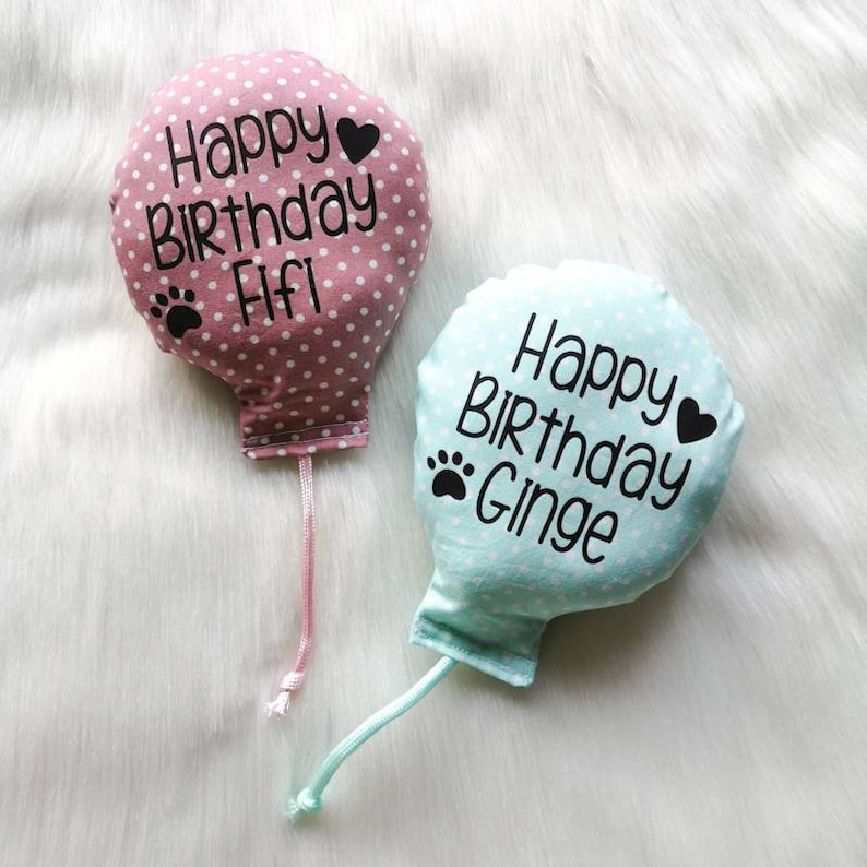 Two fabric catnip cat toy balloons with polka dots and strings. The pink balloon says 'Happy Birthday Fifi' with a paw print and a heart. The blue balloon says 'Happy Birthday Ginge' with a paw print and a heart.