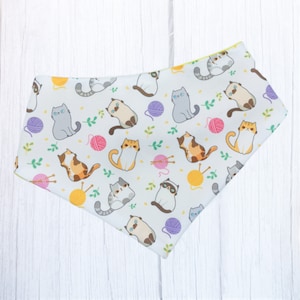 May include: White bandana with a repeating pattern of cats and yarn balls. The cats are in various poses and colors, including gray, brown, and white. The yarn balls are in shades of pink, yellow, and purple.