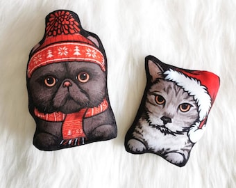 May include: Two plush cat toys wearing festive winter hats. The black cat is wearing a red and white knit hat and scarf. The gray tabby cat is wearing a red Santa hat.