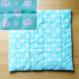 A light blue quilted cat-themed catnip pet bed with white cats and yellow flowers printed on the fabric. The bed is quilted in a grid pattern.