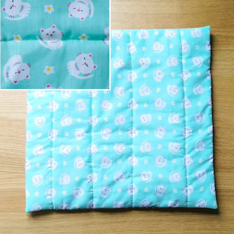 A light blue quilted cat-themed catnip pet bed with white cats and yellow flowers printed on the fabric. The bed is quilted in a grid pattern.