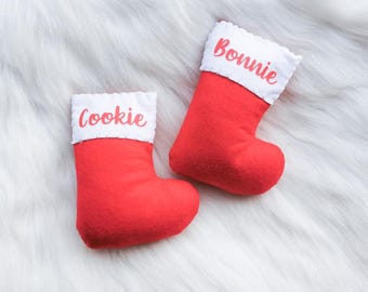 May include: Two red Christmas stockings with white cuffs. The stocking on the left has the name "Cookie" embroidered on the cuff. The stocking on the right has the name "Bonnie" embroidered on the cuff.