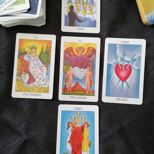 Op de afbeelding: Een spread van tarotkaarten met kleurrijke illustraties en titels als "De Keizerin", "De Geliefden" en "Drie Zwaarden". Een kaartspel en een metalen container zijn ook zichtbaar.