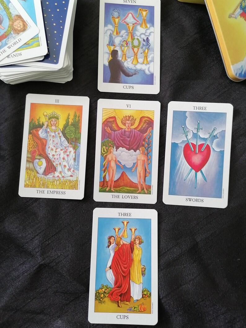 Op de afbeelding: Een spread van tarotkaarten met kleurrijke illustraties en titels als "De Keizerin", "De Geliefden" en "Drie Zwaarden". Een kaartspel en een metalen container zijn ook zichtbaar.