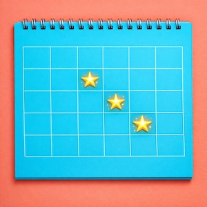 Può includere: Un calendario blu a spirale con una griglia di linee bianche. Tre icone a forma di stella dorata sono posizionate sul calendario. Lo sfondo è di colore corallo pieno. Il calendario è una rappresentazione visiva della pianificazione e dell'organizzazione.