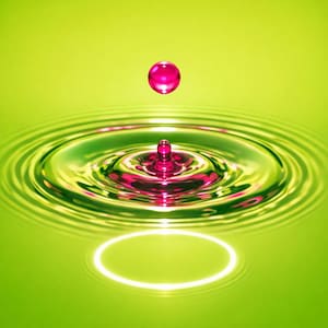 May include: A vibrant image captures a magenta droplet suspended above a pool of water, creating concentric ripples. The water and background are a bright green, with a white oval reflection at the bottom. The droplet is about to impact the water's surface.