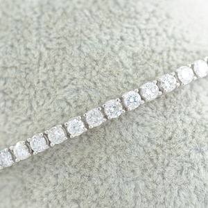 Puede incluir: Un delicado brazalete de tenis de diamantes con una fila de brillantes diamantes redondos engastados en una banda de metal blanco.