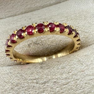 Könnte beinhalten: Ein goldener Ring mit einer durchgehenden Reihe leuchtend roter Edelsteine. Der Ring hat ein poliertes Goldband mit einem filigranen, strukturierten Muster an den Rändern. Die Edelsteine sind gleichmäßig angeordnet und bilden ein nahtloses Muster.