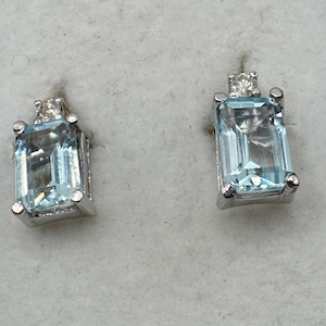 May include: A pair of silver stud earrings. Each earring features a rectangular, light blue gemstone with a smaller, clear stone at the top. The earrings have a simple, elegant design, suitable for everyday wear.