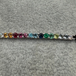 Può includere: Un braccialetto d'argento con una fila di pietre preziose multicolori, tra cui giallo, rosso, rosa, blu e verde. Il braccialetto è fissato con cordoncini neri annodati. Le pietre sono disposte in un motivo lineare.