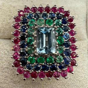 May include: A rectangular silver ring with a large, light blue rectangular gemstone at the center, surrounded by a mosaic of smaller gemstones in shades of red, green, blue, and black. The ring has a detailed, ornate design.