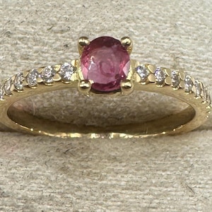 Puede incluir: Un anillo de oro con una gema rosa central y pequeñas gemas blancas a lo largo de la banda. El anillo tiene un diseño clásico y un acabado pulido. Una joya adecuada para diversas ocasiones.