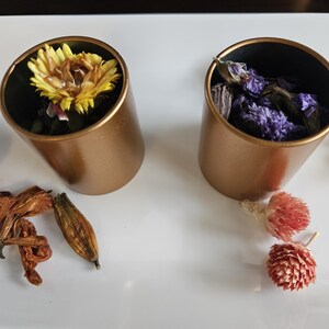 Könnte beinhalten: Zwei kupferfarbene zylindrische Behälter, jeweils gefüllt mit getrockneten Blumen. Einer enthält eine gelbe Blume, der andere lila Blumen. Verschiedene getrocknete Blumen sind um die Behälter auf einer weißen Oberfläche verstreut.