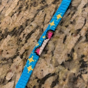 May include: A blue friendship bracelet with a white, pink, and yellow unicorn design. The bracelet is made with colorful embroidery floss.
