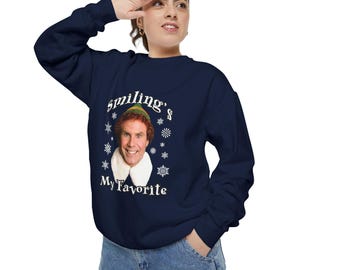 May include: Navy blue sweatshirt featuring a smiling image of a character with a white collar and green hat. The text "smiling's My Favorite" is printed above and below the image, with snowflake accents. A long-sleeved crew neck.