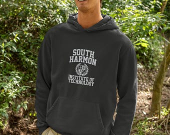May include: A dark gray hooded sweatshirt featuring the white text South Harmon Institute of Technology with a central crest emblem. The garment includes a front kangaroo pocket, long sleeves, and a hood, worn with a green baseball cap and khaki pants.
