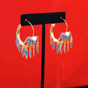 May include: Gold-tone hoop earrings with blue enamel feather-shaped accents and dangling teardrop charms. Each earring features a white pearl-like bead. The earrings are displayed against a red background.