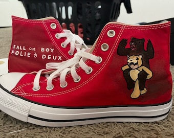 May include: Red high-top Converse sneakers with white laces and the words "Fall Out Boy Folie à Deux" painted on the side. The other side of the shoe features a hand-painted illustration of a person in a bear costume.