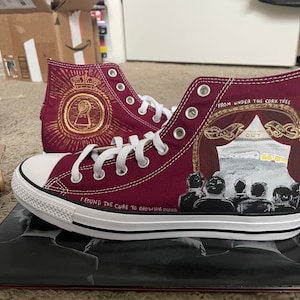 May include: Burgundy high-top sneakers with custom painted designs. One shoe features a gold keyhole design with a crown and the text "I found the cure to growing older". The other shoe features a red and gold tent with the text "From under the cork tree" and a crowd of people.