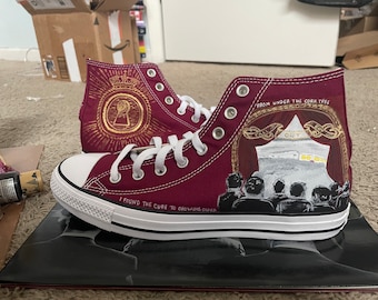 May include: Burgundy high-top sneakers with custom painted designs. One shoe features a gold keyhole design with a crown and the text "I found the cure to growing older". The other shoe features a red and gold tent with the text "From under the cork tree" and a crowd of people.