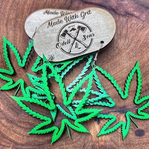 May include: Green marijuana leaf earrings made of wood. The earrings are hanging from a wooden tag that says "Made With Grit Odell & Sons".
