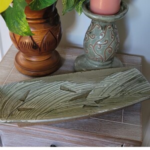 May include: A rectangular, handmade ceramic tray with a textured, abstract design in shades of green and brown. The tray has a raised edge and a series of carved lines and shapes. A wooden vase and a decorative candle holder are in the background.