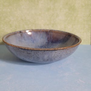 May include: A handmade ceramic bowl with a wide, shallow design. The bowl features a blend of blue and purple hues with a textured surface and a brown rim. The bowl is suitable for serving food.