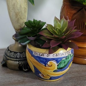 May include: A small, round, hand-painted ceramic planter featuring yellow, blue, and green scrollwork patterns with the text "INCA DI CAMASTRA" written on the rim. Two artificial succulent plants with green and purple leaves sit inside the pot.