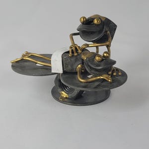 May include: A whimsical metal sculpture of two frogs. One frog is lying down, appearing to receive a massage from the other frog. The sculpture features a combination of dark gray and gold tones, with the frogs resting on circular platforms.