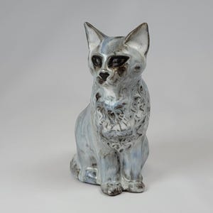 May include: A ceramic cat figurine in a seated position. The cat has a light blue and white glaze with dark brown accents around the eyes and nose. The figurine has a textured appearance.