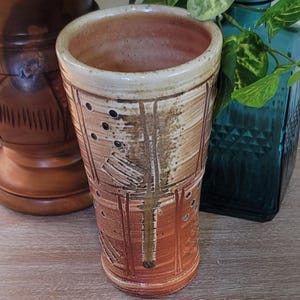 May include: A tall, cylindrical ceramic vase featuring a two-tone glaze with a creamy off-white top section and a warm terracotta-colored base. The surface is decorated with vertical incised lines, small circular indentations, and textured patterns.