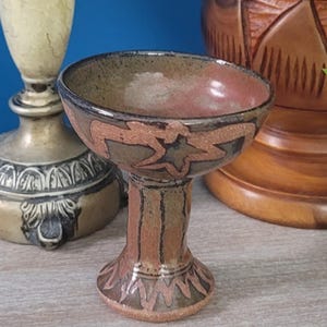 May include: A ceramic goblet featuring a wide bowl and a pedestal base. The exterior displays a mottled brown and reddish-tan glaze with dark, hand-painted star and geometric patterns encircling the bowl and the base of the stem.