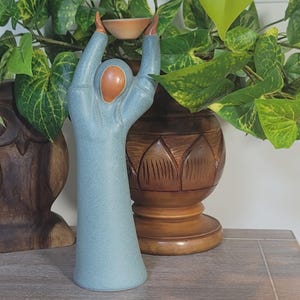May include: A tall, stylized ceramic figurine featuring a figure in a blue, speckled robe with arms raised high, holding a small, light-colored bowl above its head. The figure has a smooth, unpainted terracotta-colored face and hands.