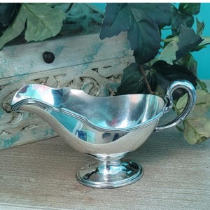 May include: A silver gravy boat with a curved spout and a handle. The boat has a pedestal base and a decorative rim. The shiny surface reflects the surrounding objects. The gravy boat is a classic serving piece.