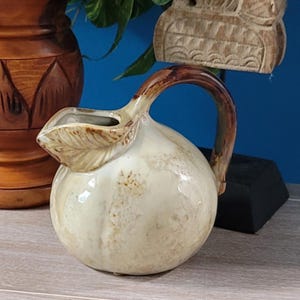 May include: A round ceramic pitcher with a glossy cream-colored glaze and brown mottled accents. The pitcher features a curved handle and a spout shaped like a textured leaf. The surface has a smooth finish with subtle variations in the glaze color.