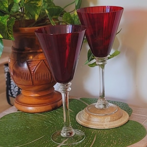 May include: Two tall wine glasses featuring deep red, conical bowls and clear glass stems with decorative ring accents at the base of the bowl. The glasses have circular clear glass bases and are positioned upright on a flat surface.