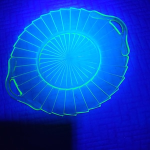 May include: A round, clear glass dish with a fluted edge and two handles. The dish glows bright green under a black light, with radial lines emanating from the center. The dish is likely vintage glassware.