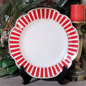 May include: A round ceramic plate with a scalloped edge featuring a white center and a border of alternating red and white vertical stripes. The rim is accented with a thin, delicate gold-colored beaded pattern along the inner edge of the stripes.