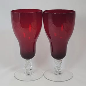 May include: Two ruby red wine glasses. The glasses have a tulip-shaped bowl and a clear, decorative stem. The base is a clear, circular foot. The glasses are made of glass and are set against a white background.