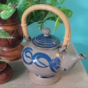 May include: A ceramic teapot with a bamboo handle and a lid. The teapot is beige with blue swirl designs and a brown lid. The handle is made of woven bamboo. The teapot is sitting on a beige base.
