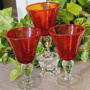 May include: Three red glass goblets with a bubbled texture and clear glass stems. The two shorter goblets have wide, flared bowls, while the taller central goblet is elevated on a clear, faceted glass base, creating a tiered arrangement.