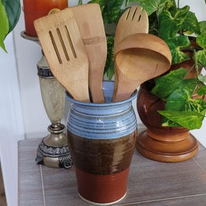 May include: A cylindrical ceramic utensil holder featuring a tri-color glaze with a light blue top, a textured dark brown middle, and a solid terracotta base. The holder contains four wooden kitchen utensils, including a slotted turner and a ladle.