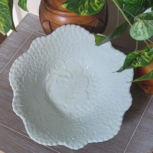 May include: A pale green ceramic bowl with a scalloped edge and a textured surface. The interior features a raised, embossed floral design in the center, surrounded by a fine, stippled pattern that covers the remainder of the bowl's surface.