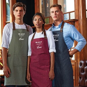May include: Three people wearing denim aprons with brown leather straps and the text "the secret ingredient is Love" embroidered on the front. The aprons are in green, red, and blue.